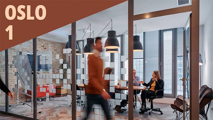 en ung mann går raskt forbi et kontor med glassvegg og to personer ved et bord