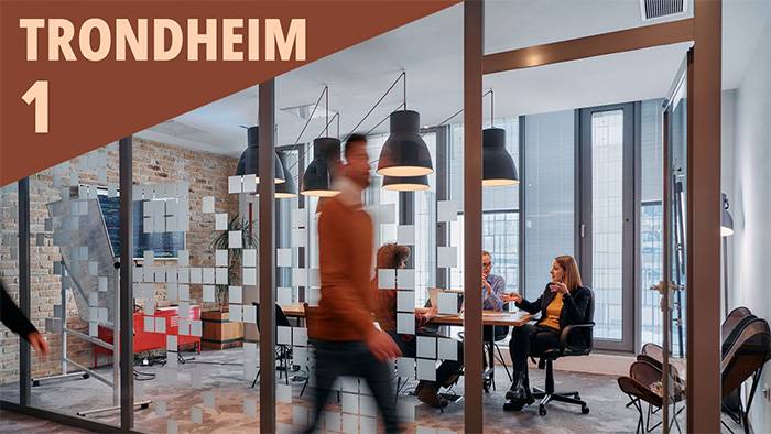 en ung mann går raskt forbi et kontor med glassvegg og to personer ved et bord